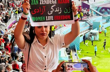 Vor den Rängen eines Fußballstadiums hält eine junge Frau in weißer Bluse ein Schild in die Kamera. Darauf steht in grün-weiß-rot: Zan Zendegi Azadi - Women Life Freedom