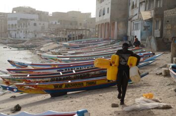 Auf dem Strand läuft ein Mann mit Wasserkanistern, im Hintergrund Holzboote, die Geflüchtete über den Seeweg aus dem Senegal wegbringen