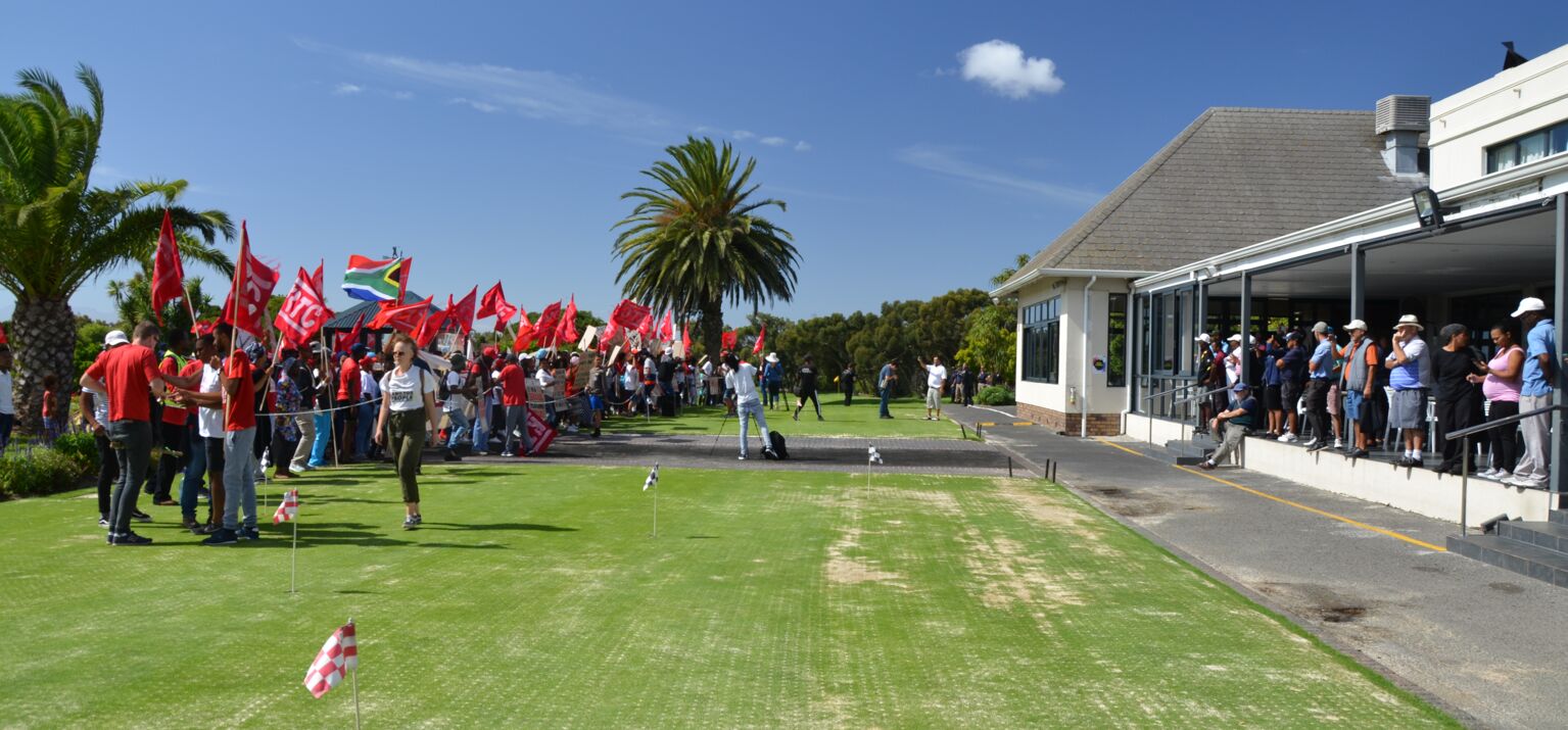 Auf dem Golfplatz Rondebosch Golf Course steht links eine größere Menschengruppe mit roten Fahnen, rechts stehen Golfer*innen unter einem Dach.