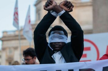 mit verbundene Augen und erhobenen Händen steht Y. David hinter einem Transparent aur der Demonstration gegen italienische Grenzpolitik