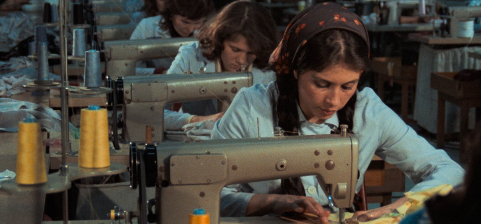 Filmstill aus »Kara Kafa«. Frauen in blauen Uniformen sitzen in einer Reihe über Nähmaschinen gebeugt.