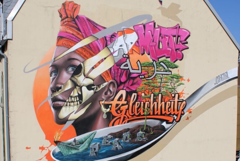 Ein Graffiti an einer Hauswand, das eine junge afrikanische Frau darstellt, sowie die entwicklungspolitischen Schlagwörter "Armut", Gleichheit" und "Peace".
