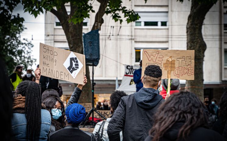 Bei der Mahnwache im Gedenken an George Floydin Freiburg, 06. Juni 2020 sind Menschen zu sehen, meist von hinten. Zwei Schilder sind zu lesen: Stop Killing The Mandem und We For The Change. Foto: Sévérine Kpoti