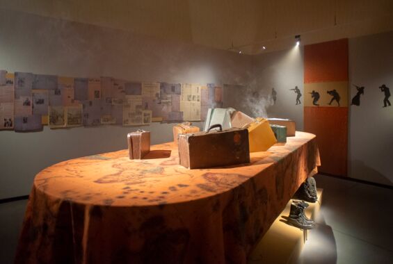 Ein Tisch mit einer Tischdecke mit der Landkarte von Afrika und Koffern darauf steht in einem Ausstellungsraum. Über ihm steigt Rauch auf.