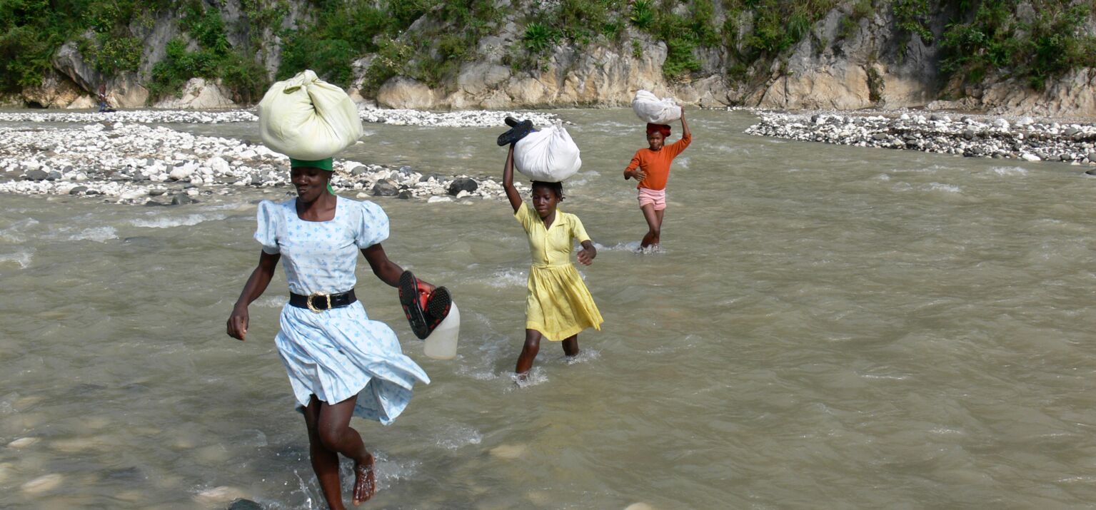 Eine Frau und zwei Kinder mit Büneln auf dem Kopf durchqueren einen Fluss