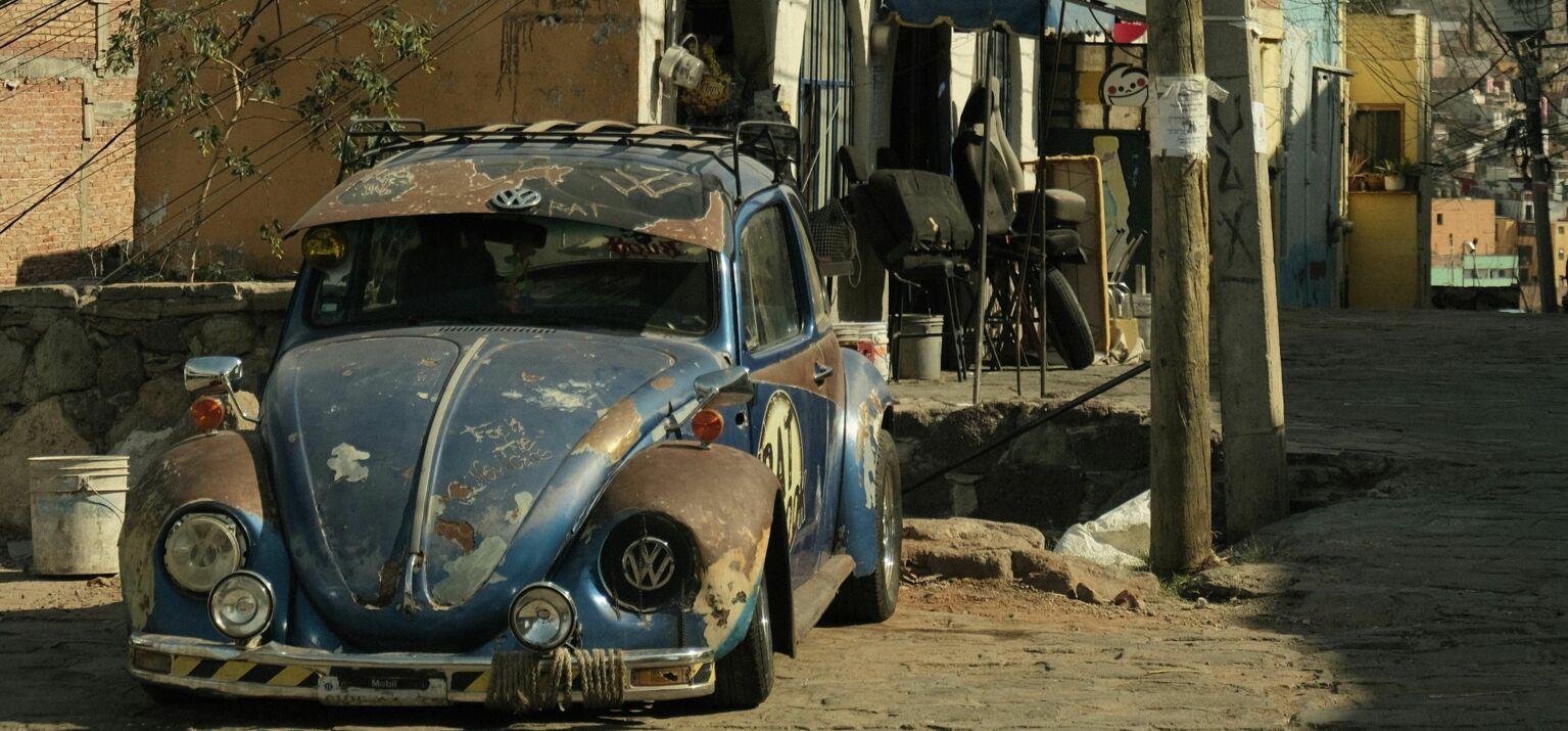 Ein liegen gebliebener, kaputter VW Käfer vor heruntergekommenen Häusern, Berge im Hintergrund. Krisen nehmen zu.