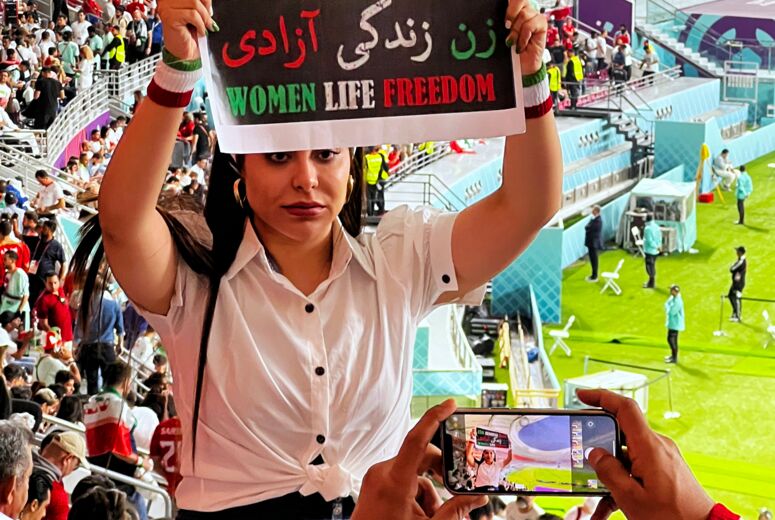 Vor den Rängen eines Fußballstadiums hält eine junge Frau in weißer Bluse ein Schild in die Kamera. Darauf steht in grün-weiß-rot: Zan Zendegi Azadi - Women Life Freedom