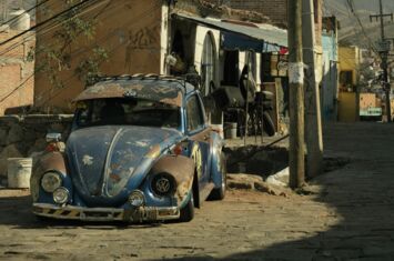 Ein liegen gebliebener, kaputter VW Käfer vor heruntergekommenen Häusern, Berge im Hintergrund. Krisen nehmen zu.