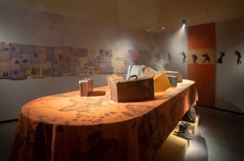 Ein Tisch mit einer Tischdecke mit der Landkarte von Afrika und Koffern darauf steht in einem Ausstellungsraum. Über ihm steigt Rauch auf.