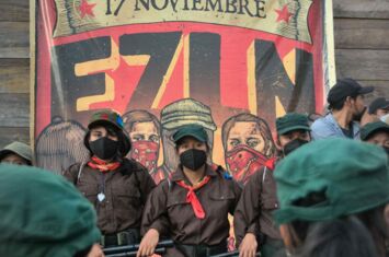 Vermummte EZLN-Kämpfer*innen bei den Jubiläumsfeiern vor einem Plakat in Chiapas