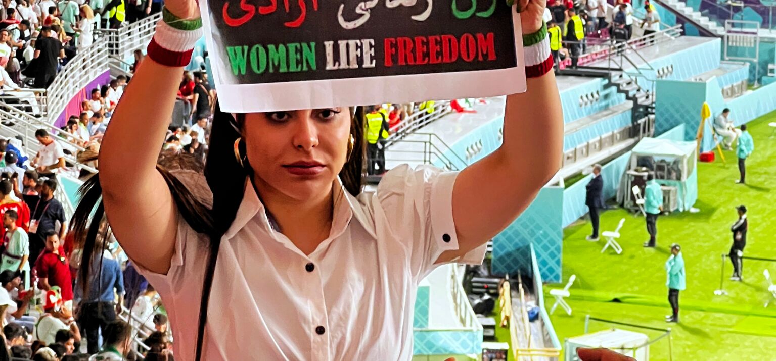 Vor den Rängen eines Fußballstadiums hält eine junge Frau in weißer Bluse ein Schild in die Kamera. Darauf steht in grün-weiß-rot: Zan Zendegi Azadi - Women Life Freedom