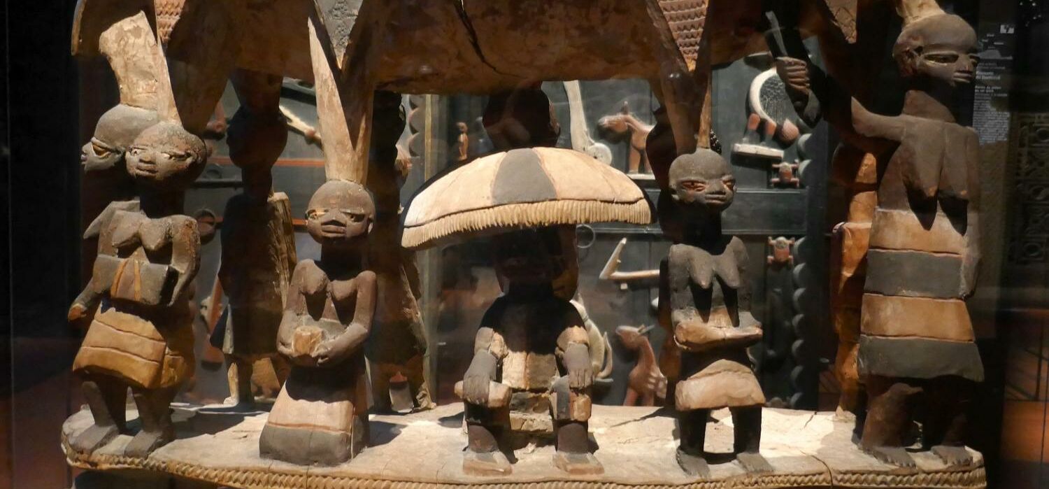 Aus Holz geschnitzte Miniaturfiguren - Königssitz mit mit Figuren der Yoruba-Kultur, ausgestellt in einer Vitrine