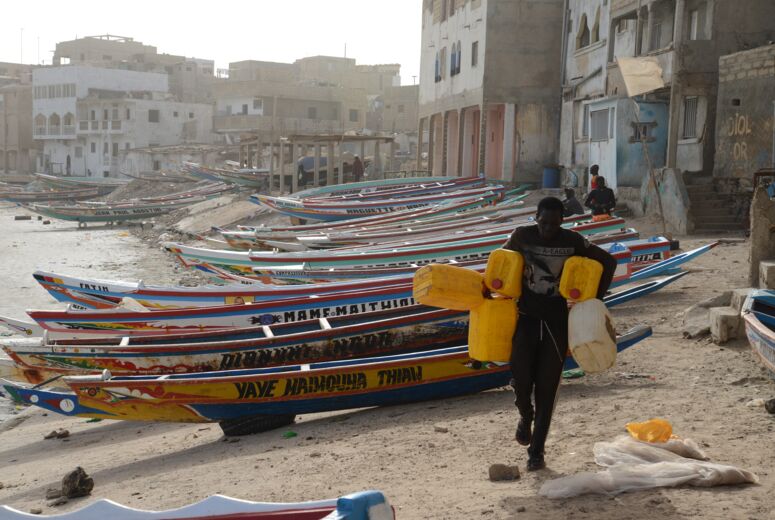 Auf dem Strand läuft ein Mann mit Wasserkanistern, im Hintergrund Holzboote, die Geflüchtete über den Seeweg aus dem Senegal wegbringen